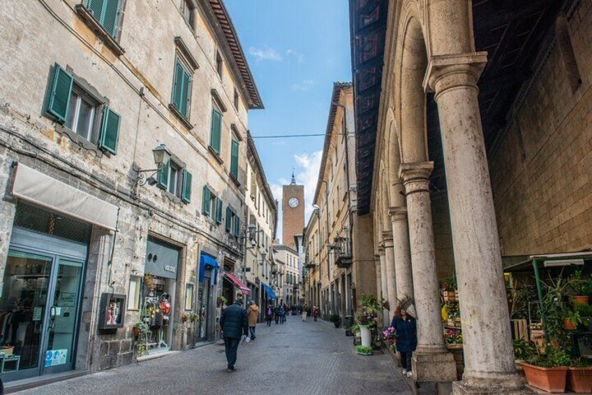 A narrow street with historic buildings, a clock tower, and pedestrians. 3