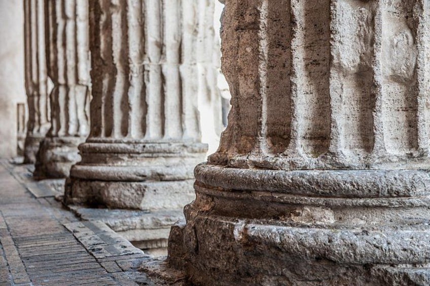 A row of weathered stone columns with fluted shafts and capitals, set in a straight line on a paved surface. 3