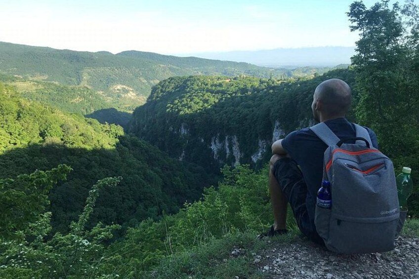 Eine Person sitzt auf einer Felsvorsprung und blickt auf ein üppiges Tal mit dichtem Wald und einer steilen Klippe. 4