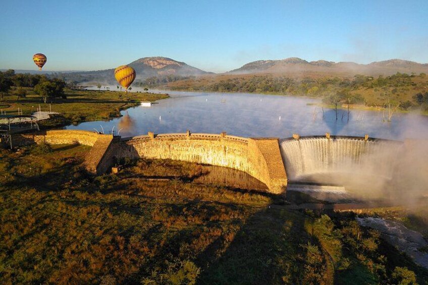 A dam with water flowing over it, hot air balloons in the background, and a hilly landscape. 4