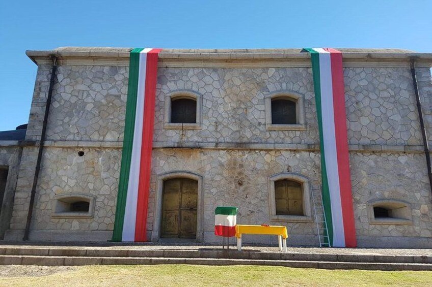 A stone building with a flag on top and a bench in front. 2