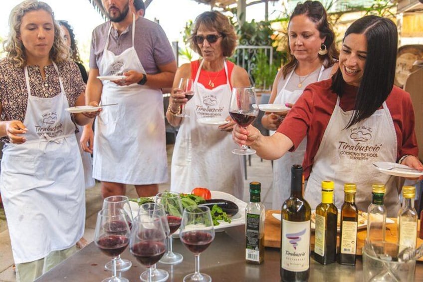 Un grupo de personas con delantales en un evento de cocina, sosteniendo platos y copas de vino, con botellas de vino y una ensalada en la mesa. 3