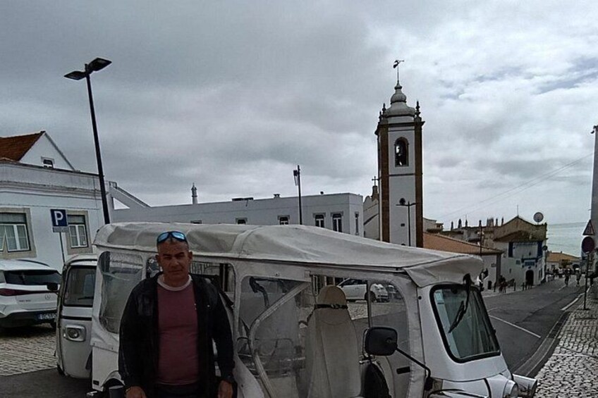 A man standing in front of a white van with a tent on top, a church with a bell tower, and a paved street with buildings. 4
