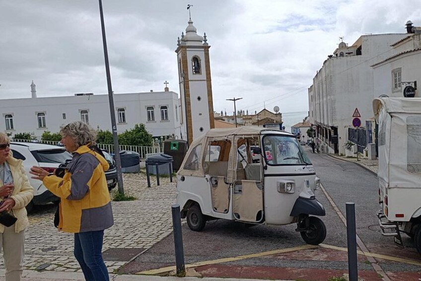 A street scene with a small white vehicle, a church tower, and a building with a clock. 3