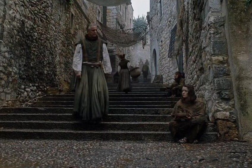 A medieval street with stone buildings, a person walking up stairs, and another person sitting on the steps. 4