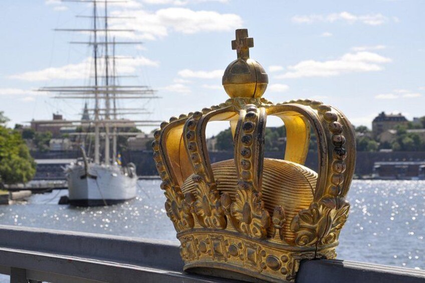 Una corona dorada con diseños intrincados, un barco con varios mástiles al fondo y una masa de agua que refleja el cielo. 2