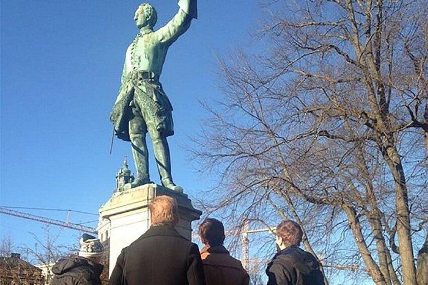 Una estatua de bronce de un hombre con el brazo levantado, un pedestal y árboles sin hojas al fondo. 1