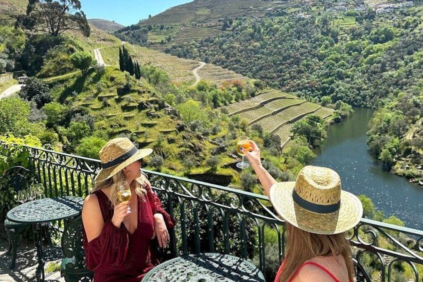 Zwei Personen genießen einen Drink auf einer Terrasse mit Blick auf einen Fluss und terrassenförmig angelegte Weinberge. 1