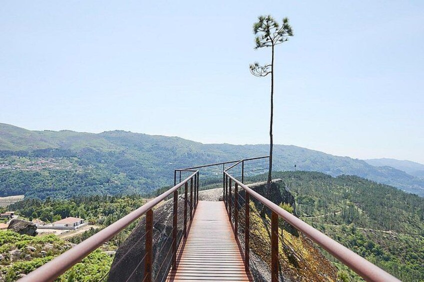 Eine Holzbrücke mit Metallgeländern, die zu einer Aussichtsplattform über einer gebirgigen Landschaft führt. 2