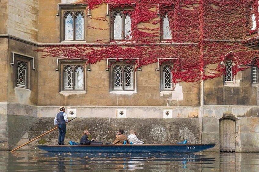Un bateau à rames avec trois personnes à bord : un homme qui rame et deux passagers, devant un bâtiment orné de lierre rouge et doté d’arcs en plein cintre. 4