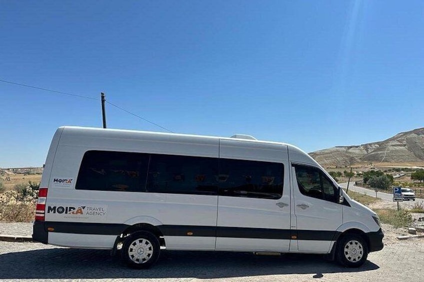 Una camioneta blanca de Moira Travel Agency estacionada en una carretera, con cielo despejado y un paisaje montañoso al fondo. 3
