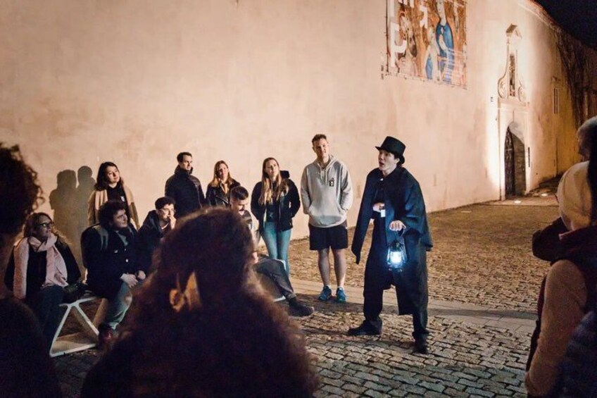 Un groupe de personnes s’est rassemblé dans un espace extérieur, sur une place pavée de pierres, devant un bâtiment orné d’une fresque murale, et une personne tenait une lampe de poche. 4