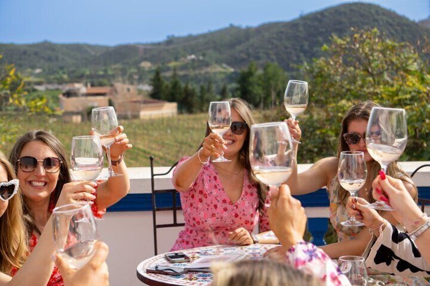 Un groupe de personnes trinquant avec des verres de vin blanc autour d’une table en plein air, avec une colline pittoresque en arrière-plan. 1