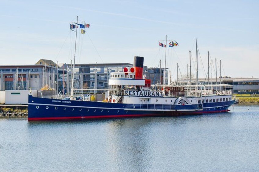 A blue and white river cruise ship named 'Pension Gloria' docked at a pier with flags, buildings, and a clear sky in the background. 3
