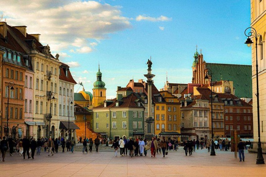 A bustling square with historic buildings, a prominent green-domed tower, and a statue in the center. 1