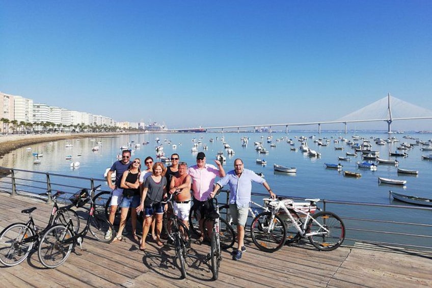 A group of people on a wooden boardwalk by a body of water with boats, a bridge, and buildings in the background. 1