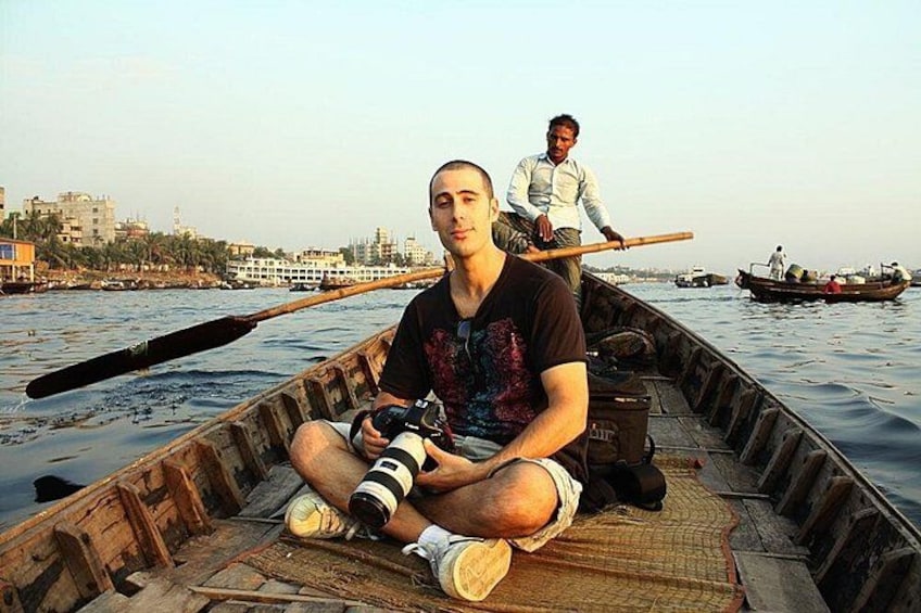 A man sitting in a wooden boat on a river, holding a camera. Another person stands behind him. 4