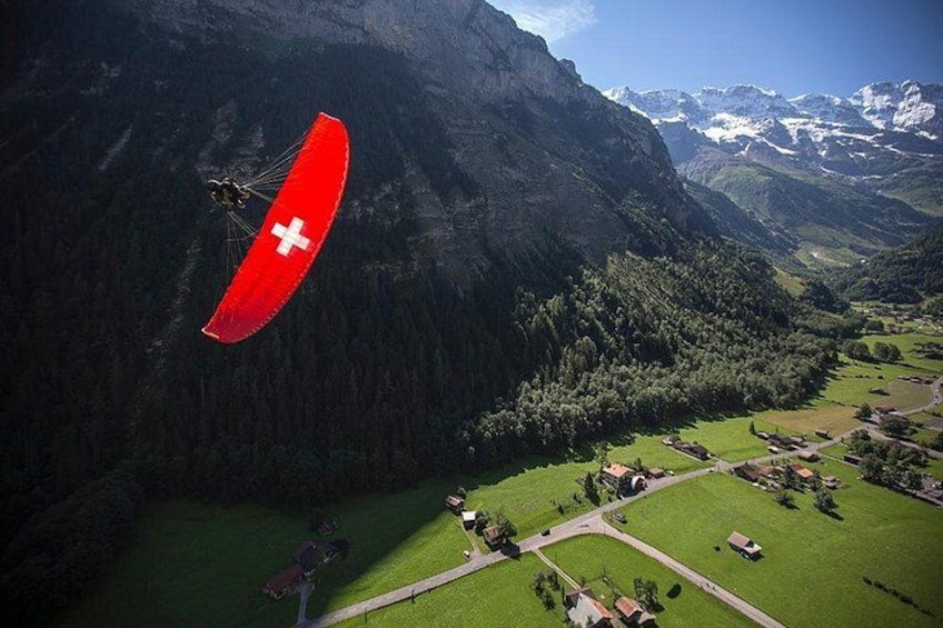 A paraglider with a red and white flag flying over a mountainous landscape with green fields and scattered buildings. 3