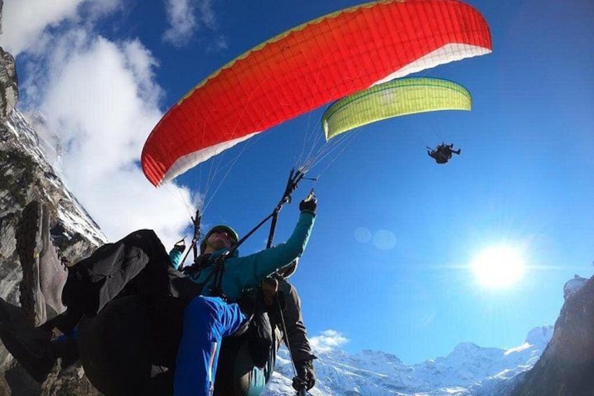 Una persona haciendo parapente con un paracaídas rojo y amarillo, cielo azul despejado y montañas nevadas al fondo. 1