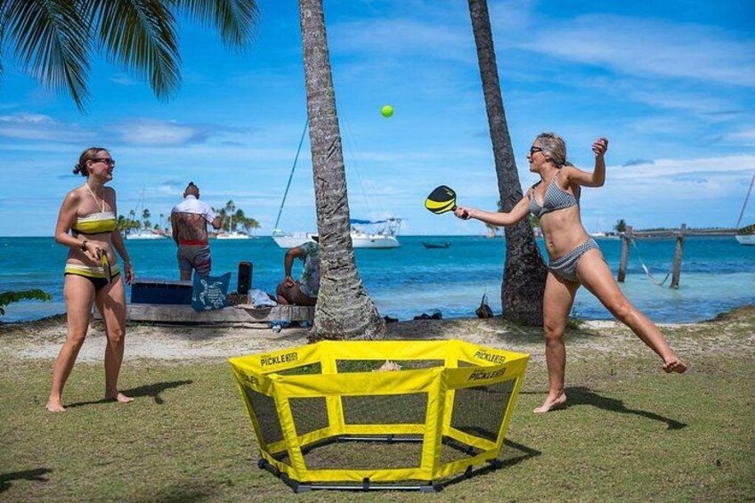 Twee personen spelen strandvolleybal bij een geel net, met palmbomen en boten op de achtergrond. 3