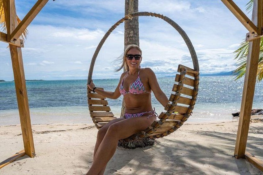 Une personne est assise dans un fauteuil à bascule en bois sur une plage de sable, avec un ciel bleu dégagé et une mer calme en arrière-plan. 4
