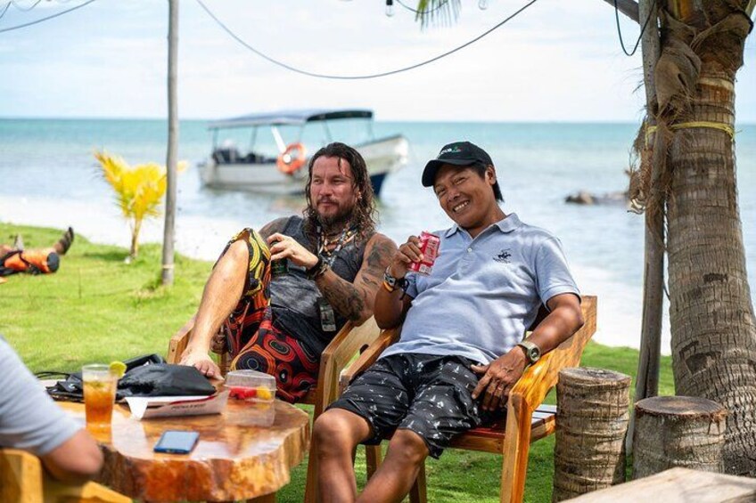 Two people sitting on a wooden chair by a palm tree, with a boat in the background and a grassy area with a small plant. 2