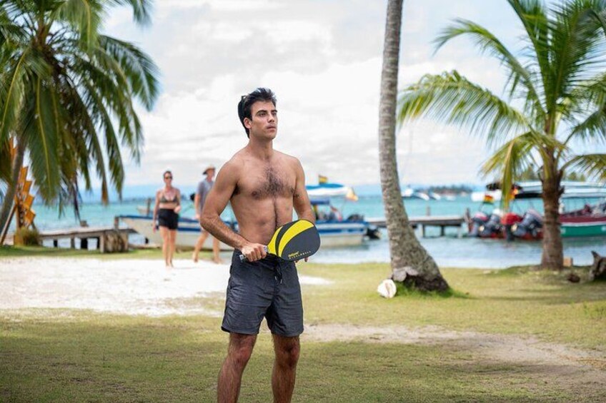 A person holding a yellow and black frisbee on a beach with palm trees and boats in the background. 3