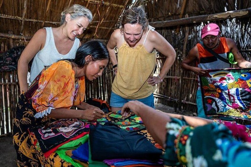 People working on colorful fabric, possibly creating traditional garments, in a rustic setting with a thatched roof. 1