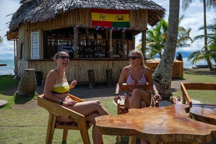 Two people sitting at wooden tables outdoors with a thatched-roof structure in the background. 5