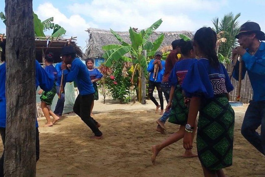 A group of people in blue shirts performing a dance in a sandy outdoor setting with banana trees and a thatched-roof structure in the background. 1