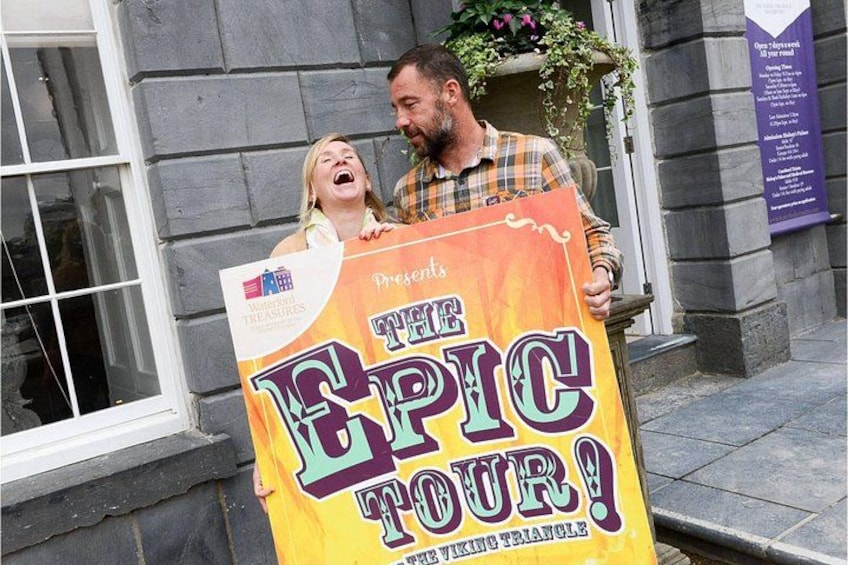 Una persona sostiene un cartel grande que dice 'THE EPIC TOUR!' con un logotipo y texto que indica un evento de búsqueda del tesoro. 1