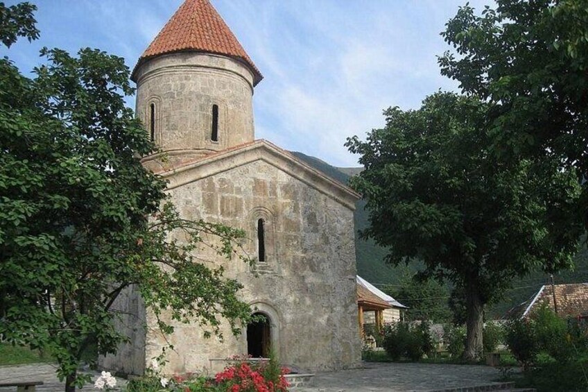 A stone church with a red-tiled roof, surrounded by trees and a clear sky. 2