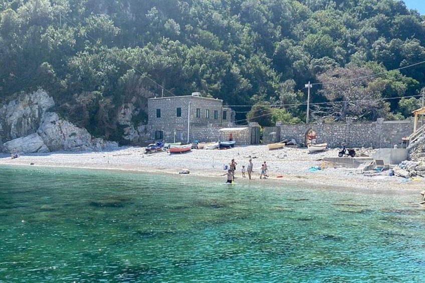 A coastal scene with a clear blue sea, a rocky beach, a stone building, and boats. 3