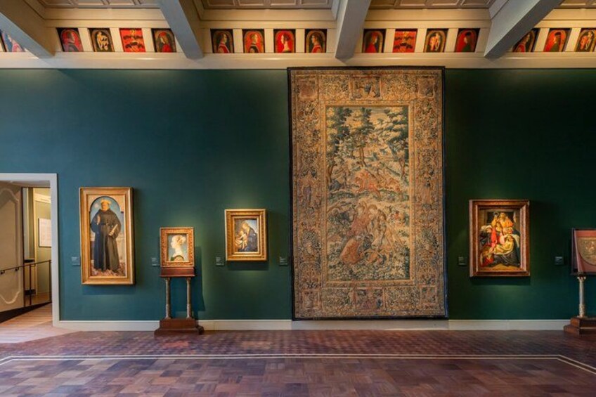 Ett galleri med inramade konstverk på turkosa väggar, en stor väv och ett trägolv. 2