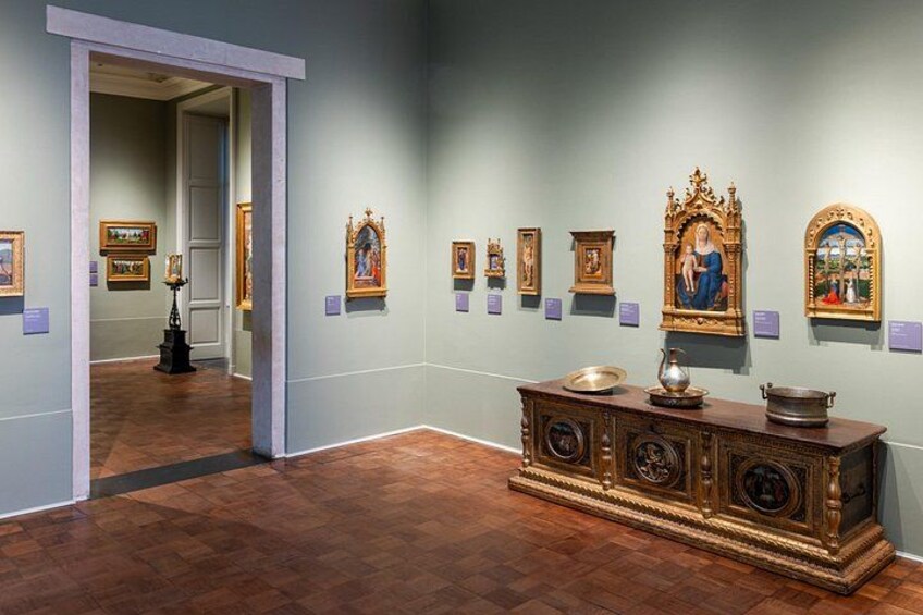 Ett konstgalleri med trägolv och ljusblå väggar. Här finns flera inramade målningar och ett stort, utsmyckat skåp med en skål och en kanna ovanpå. 3