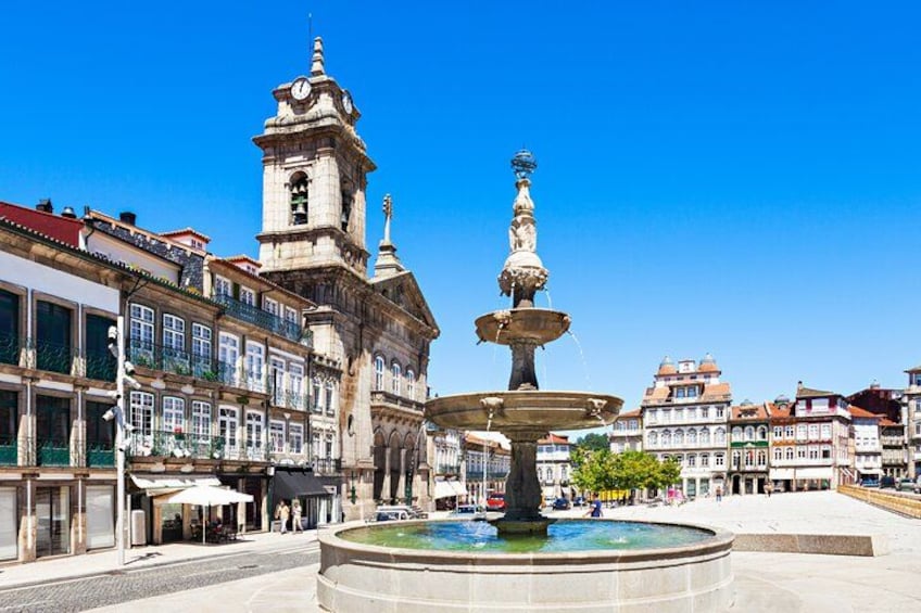 A historic square with a prominent clock tower, a central fountain, and surrounding classical architecture. 5