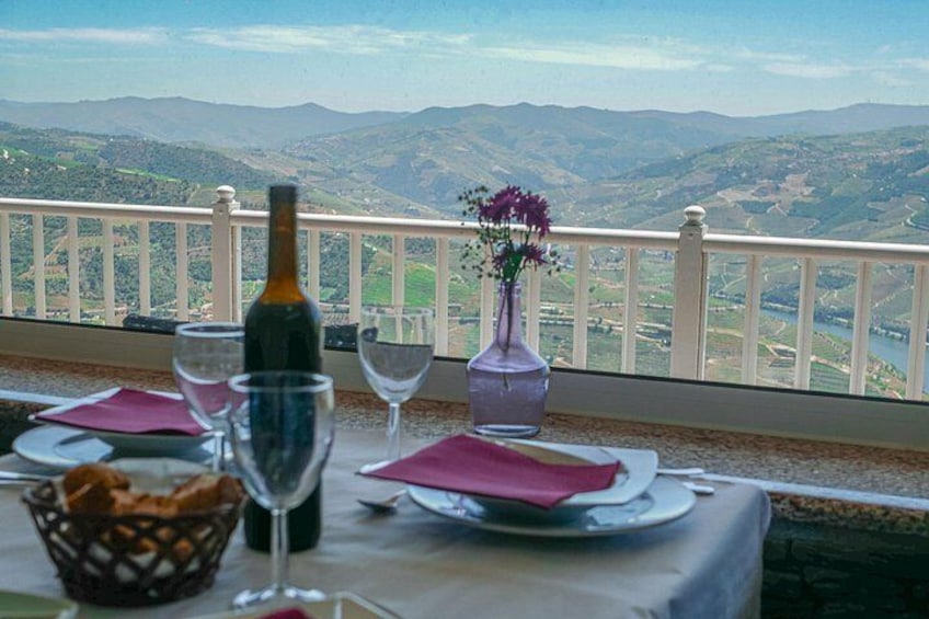 Una mesa para dos con una botella de vino, dos copas de vino, un jarrón con flores y una cesta de pan en un balcón con vistas a un paisaje montañoso. 2