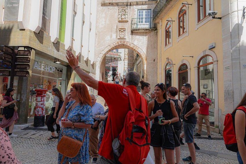Un groupe de personnes se tient sur une rue pavée, dans le centre historique d’une ville, entouré de bâtiments dotés de fenêtres en arc de cercle et d’une arche imposante. 3