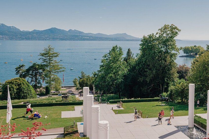 Un parc au bord du lac où l’on peut voir des gens se promener, une statue, ainsi qu’une vue sur le lac et les montagnes. 2