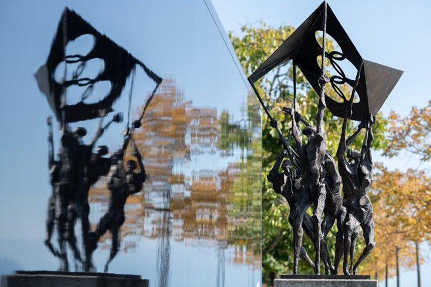 Une sculpture représentant des personnages tenant un drapeau, dont le reflet se mire dans une étendue d’eau. 4
