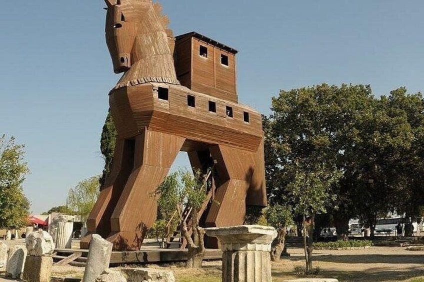 A large wooden horse sculpture with a building on its back, set in a park with trees and stone structures. 1