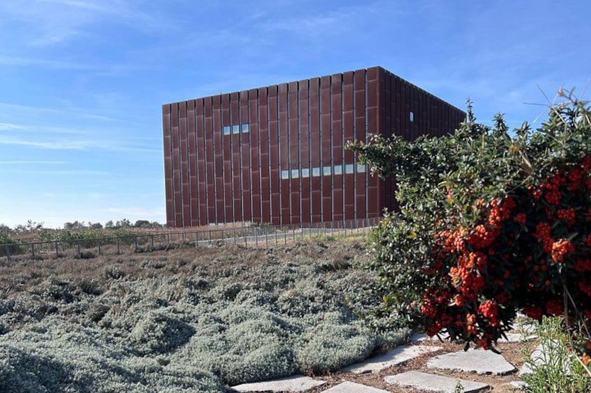 A modern building with a red facade, surrounded by a dry, grassy landscape with scattered bushes and a flowering plant. 2