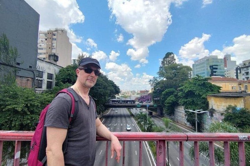Una persona parada sobre un puente, con una mochila, observando una carretera con vehículos y edificios al fondo. 3