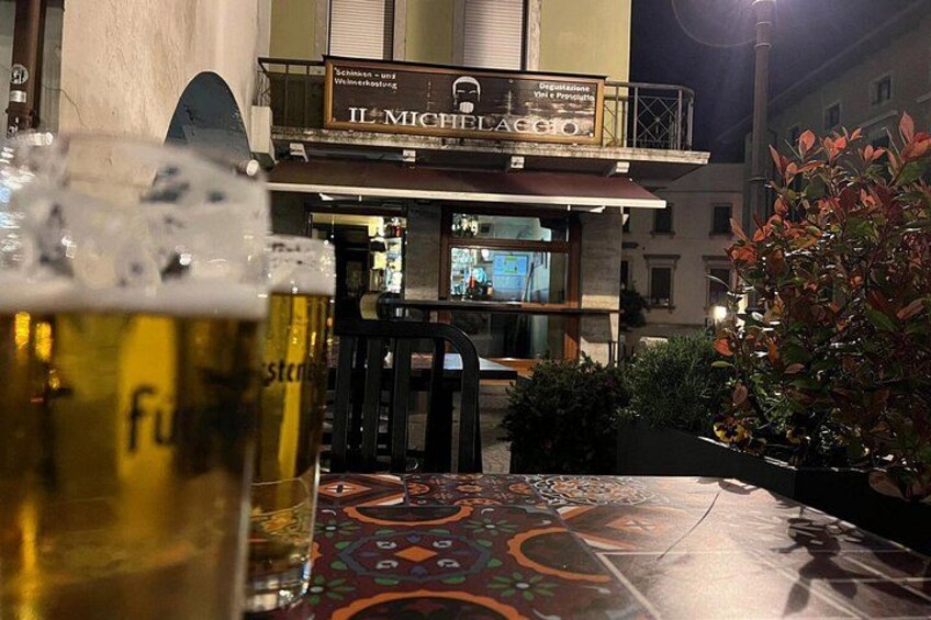 Ein Tisch mit einem Bierglas und einer Tischdecke, ein Gebäude mit einem Schild, auf dem 'Il Micheli' steht, und ein Balkon mit Geländer. 3