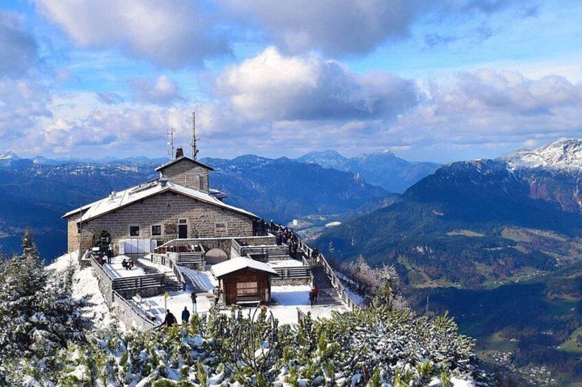 Un refugio de montaña con techo cubierto de nieve, rodeado de árboles cargados de nieve y una vista panorámica de montañas distantes. 5