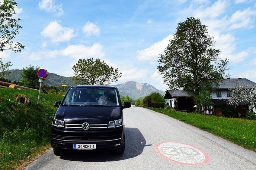 A black Volkswagen van parked on a rural road with a speed limit sign, a red circular marking on the road, and houses with trees in the background. 3