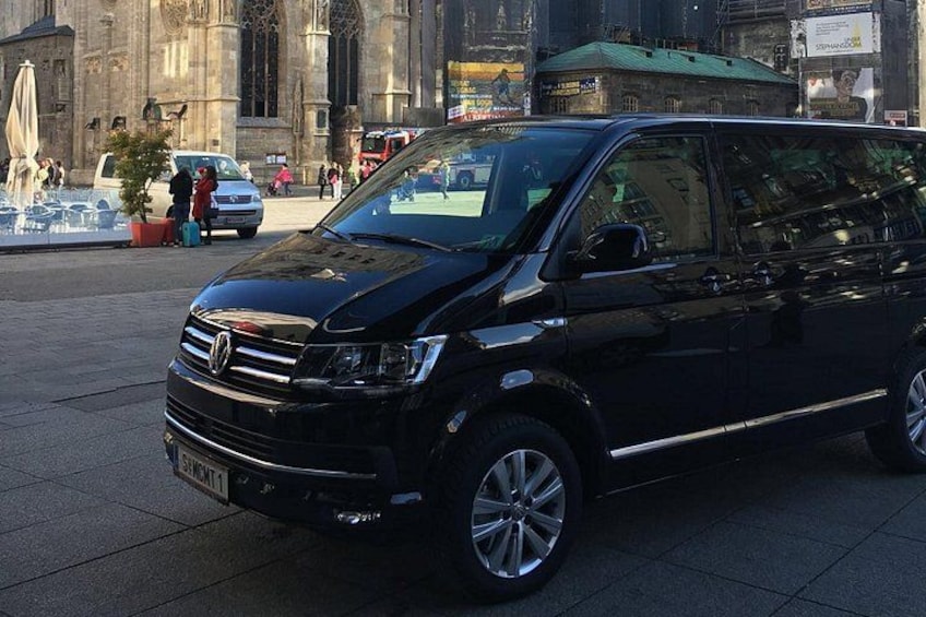A black van parked on a city street with buildings and pedestrians in the background. 2