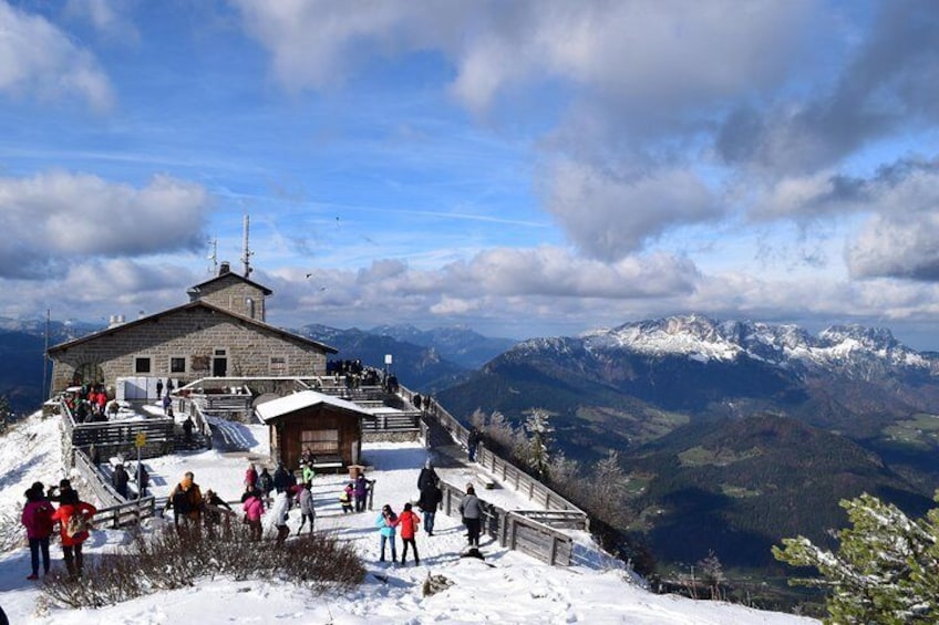 Un rifugio alpino con una baita in legno e un edificio in pietra, circondato da montagne innevate e un cielo sereno. 1