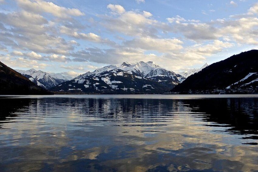 A calm lake reflecting mountains with snow-capped peaks under a partly cloudy sky. 1