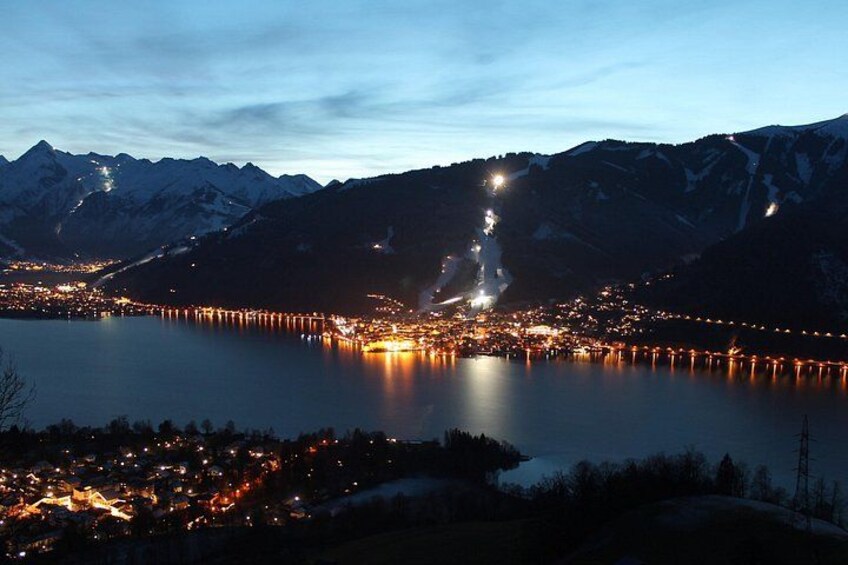 A nighttime view of a lake with a town on the shore, mountains in the background, and lights reflecting on the water. 5